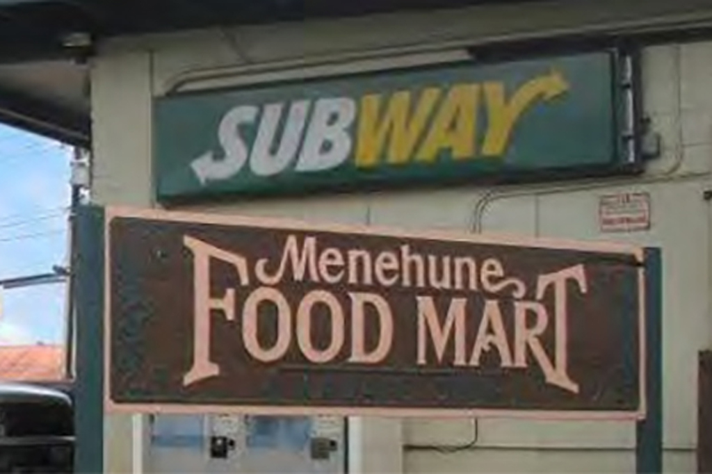 The sign for the Menehune food mart, and a subways sign above it on a beige wall.