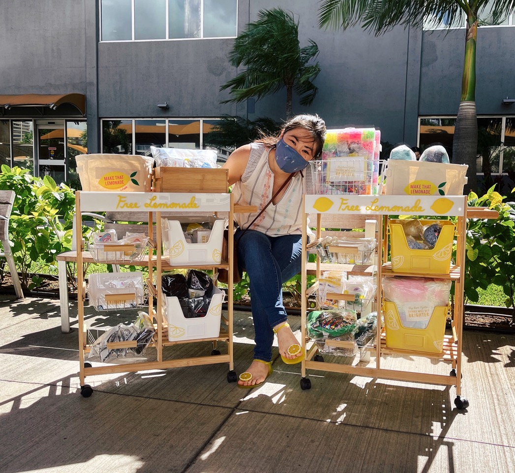 Seated woman wearing face mask posing next to carts with fund-raising food items